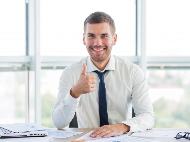 Smiling man giving thumbs up behind desk illustrates blog "5 Reasons To Use a Tax Professional"