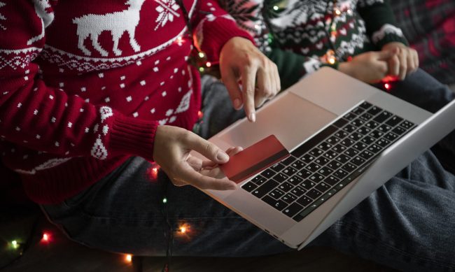 laptop-holiday-fp Couple wearing Christmas sweaters and holding credit card with laptop in their lap illustrates blog "What You Need To Know Before Donating to a Charity This Holiday Season"